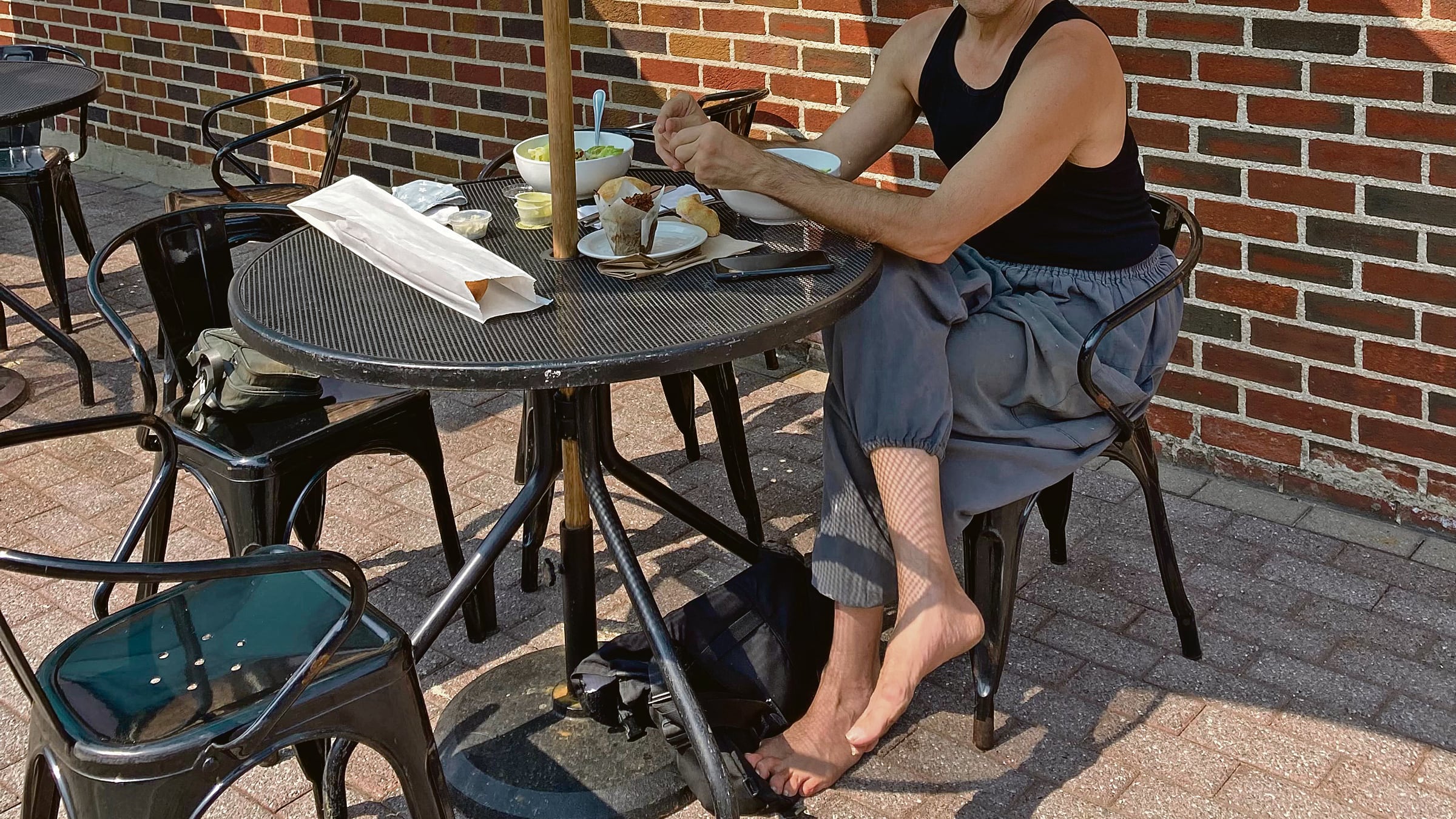 En una mesa de un establecimiento de comida, al exterior, Joseph DeRuvo está sentado, con comida en la mesa, en camisilla negra, pantalón gris, y descalzo.